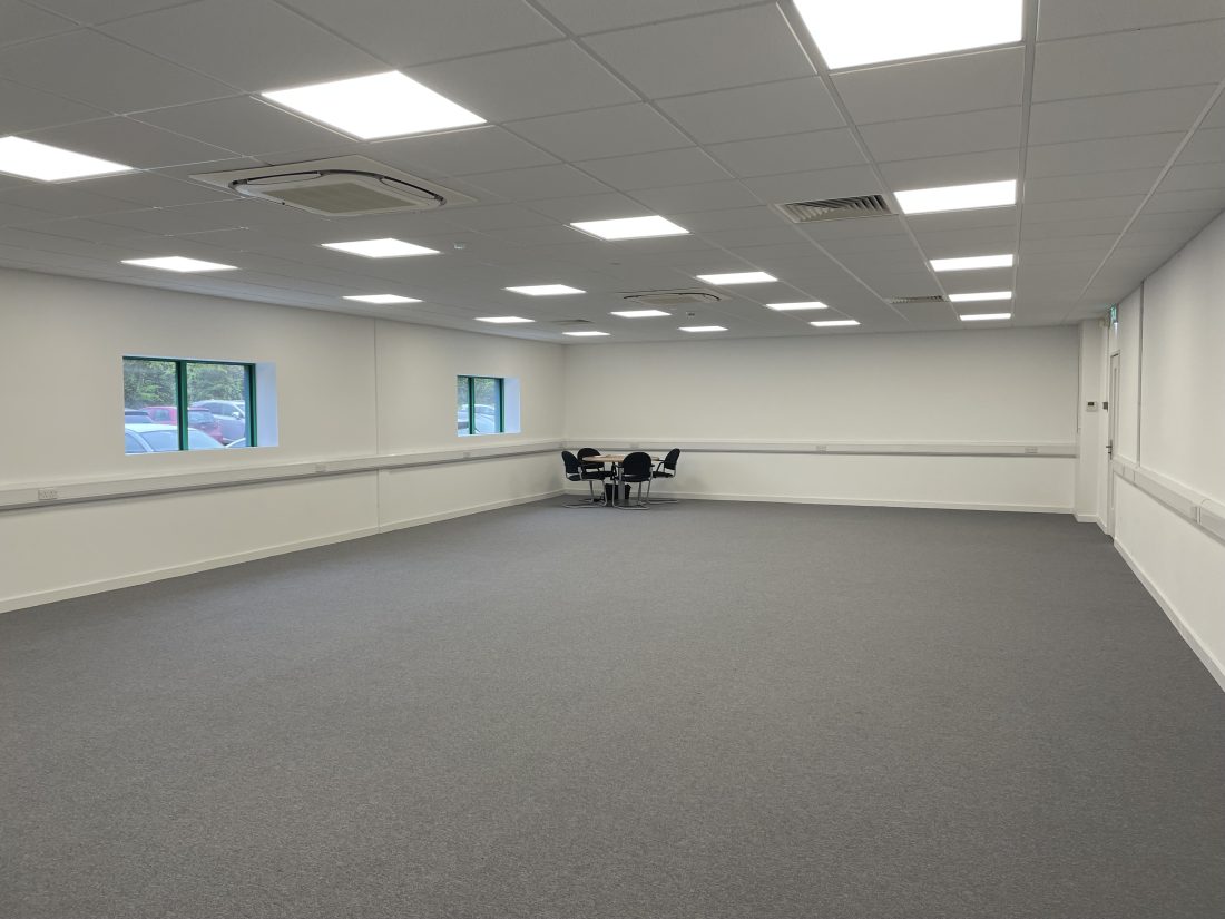 A large vacant office space, grey carpet, ceiling tiles and white walls